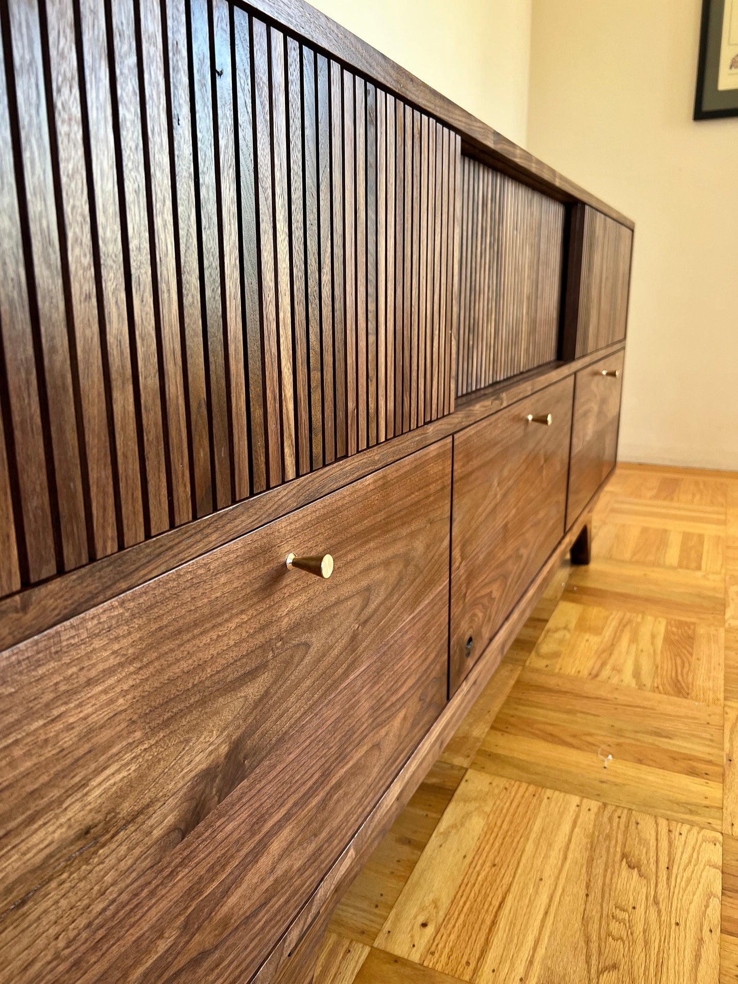 A close up photo of rectangular slat texture of the cabinets sliding doors. 3 drawers line the bottom, with even reveals around the drawers perimeter. About 2 inches from the top and centered in each drawer is a tapering, brass finger pull.
