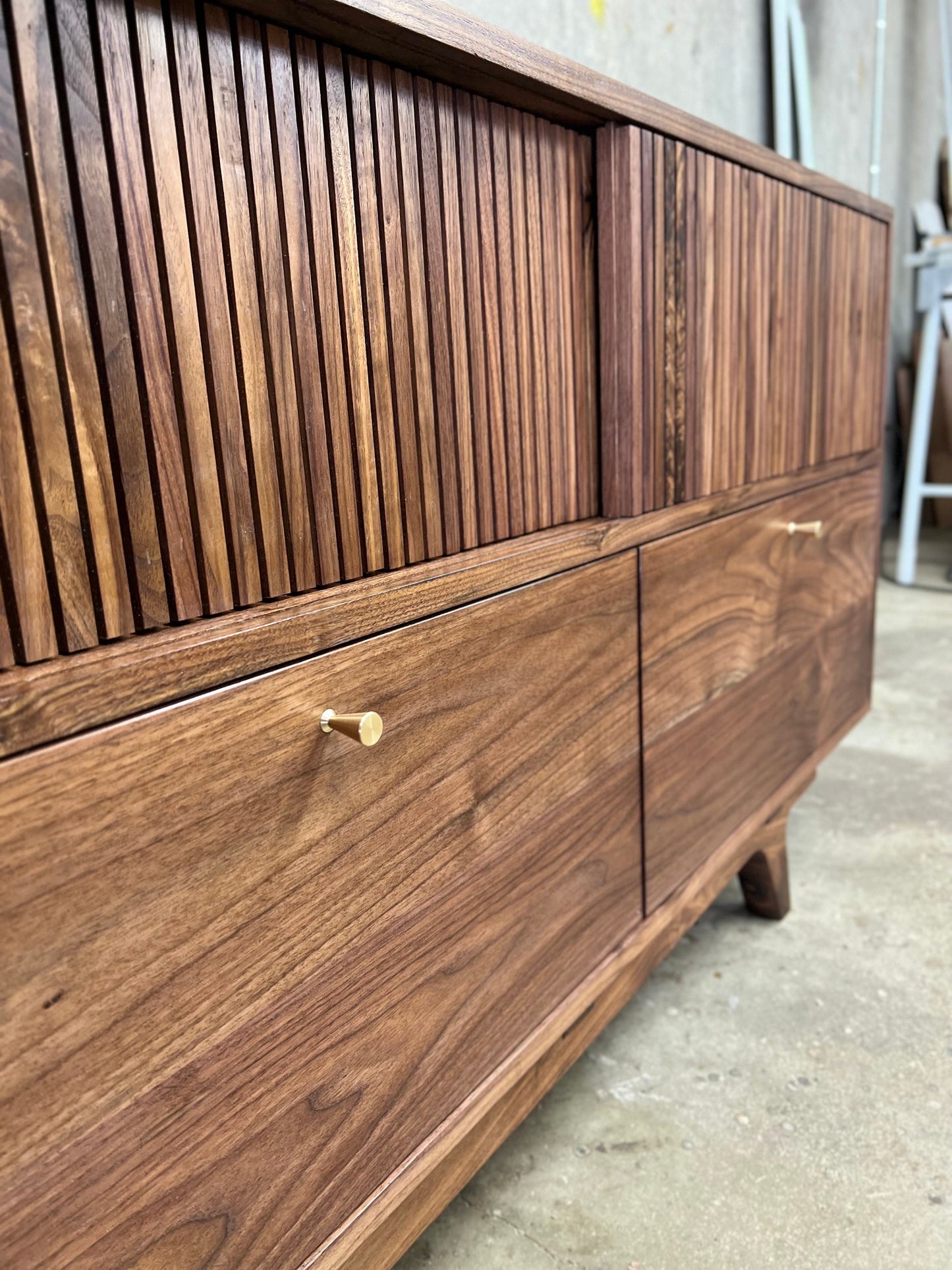 A close up photo of rectangular slat texture of the cabinets sliding doors. 3 drawers line the bottom, with even reveals around the drawers perimeter. About 2 inches from the top and centered in each drawer is a tapering, brass finger pull.